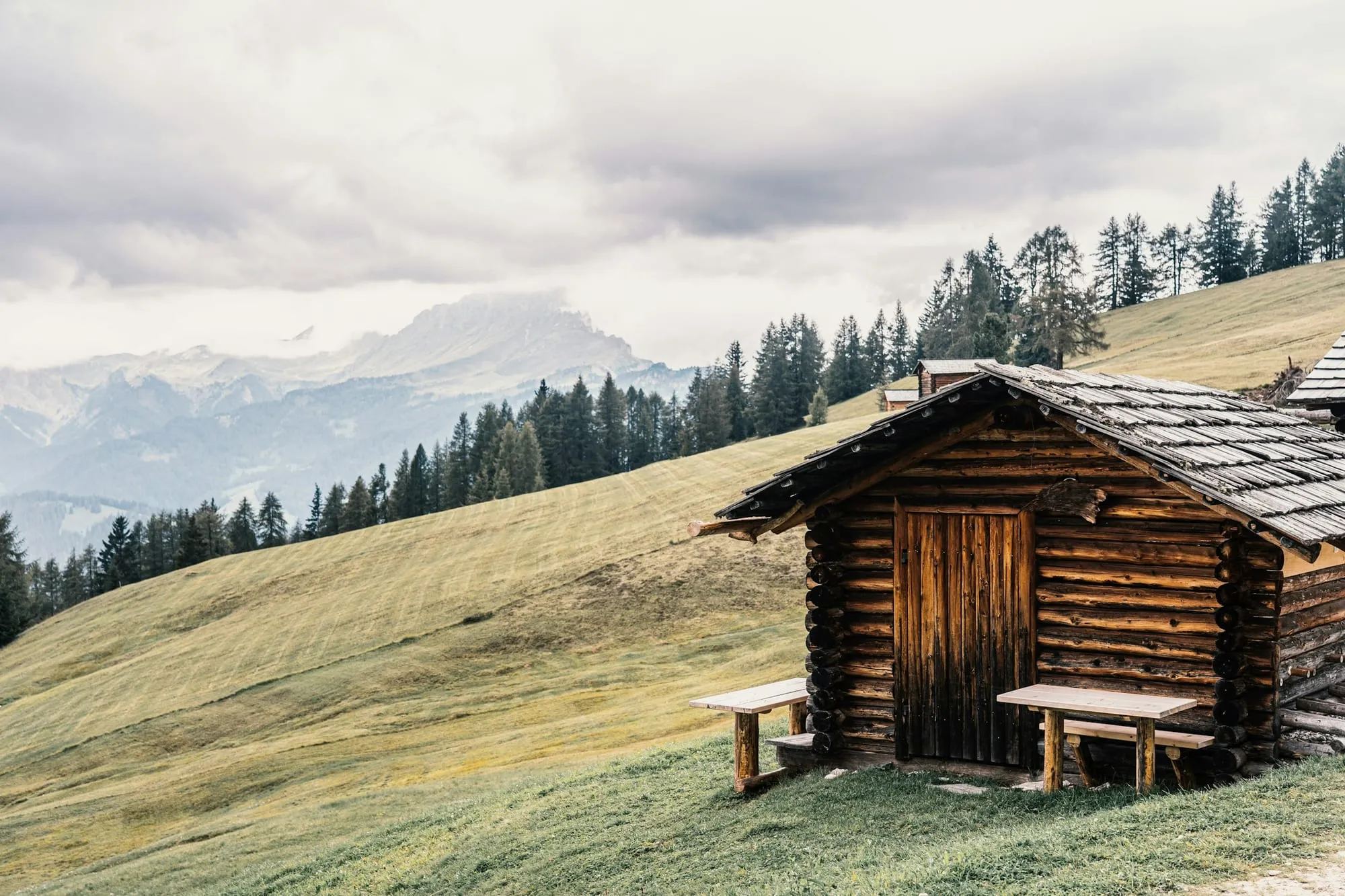 Blockhütte mit zwei Holzbänken an einem grünen Berghang, dahinter Wald und Gipfel im Dunst