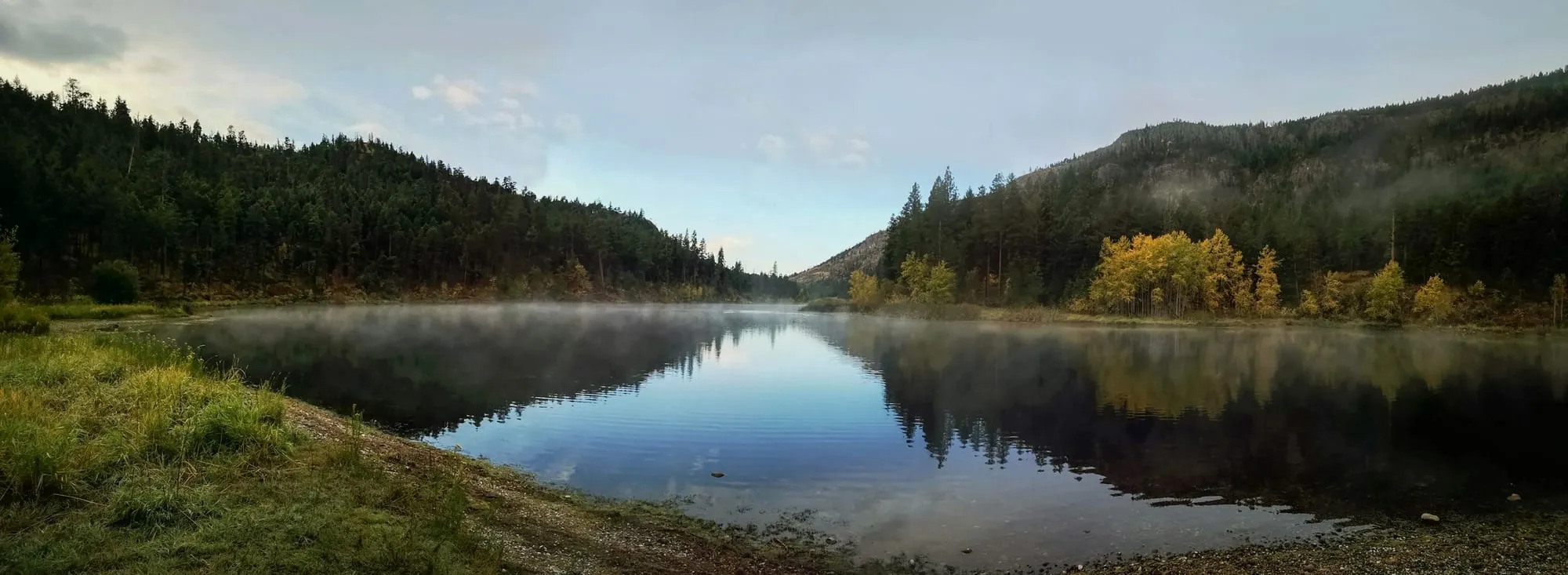 Bergsee zwischen bewaldeten Hängen, im Wasser spiegeln sich Bäume und Himmel
